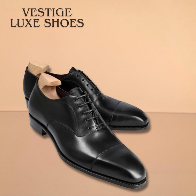 Puede incluir: Un par de zapatos de vestir de cuero negro con hormas de madera. Los zapatos tienen un dise&ntilde;o cl&aacute;sico de puntera y est&aacute;n atados con cordones. El texto "VESTIGE LUXE SHOES" est&aacute; en la parte superior de la imagen.