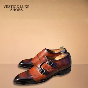 May include: A pair of brown leather double monk strap shoes with a dark brown toe cap. The shoes have silver buckles and the text "VESTIGE LUXE SHOES" is in the upper left corner.