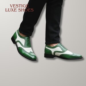 May include: A pair of two-tone green and white wingtip oxford shoes with lace-up closures and decorative brogue detailing. The text VESTIGE LUXE SHOES is visible in the upper left corner. The shoes are worn with black trousers.