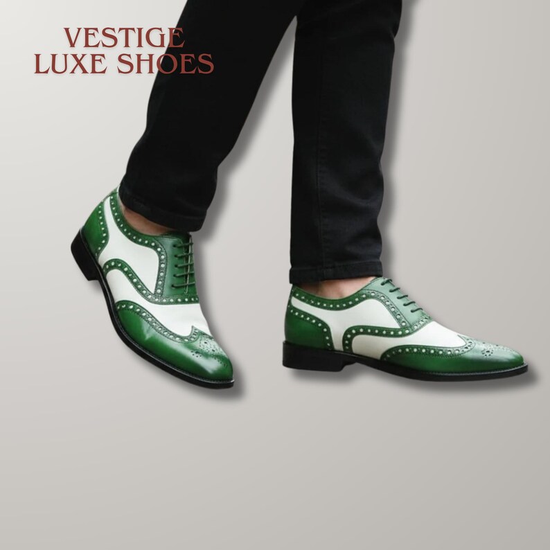 May include: A pair of two-tone green and white wingtip oxford shoes with lace-up closures and decorative brogue detailing. The text VESTIGE LUXE SHOES is visible in the upper left corner. The shoes are worn with black trousers.