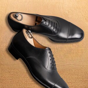 Puede incluir: Dos zapatos de vestir de cuero negro con hormas de madera. Los zapatos est&aacute;n atados con cordones y tienen un dise&ntilde;o de puntera. El texto "VESTIGE LUXE SHOES" est&aacute; en la parte superior de la imagen.