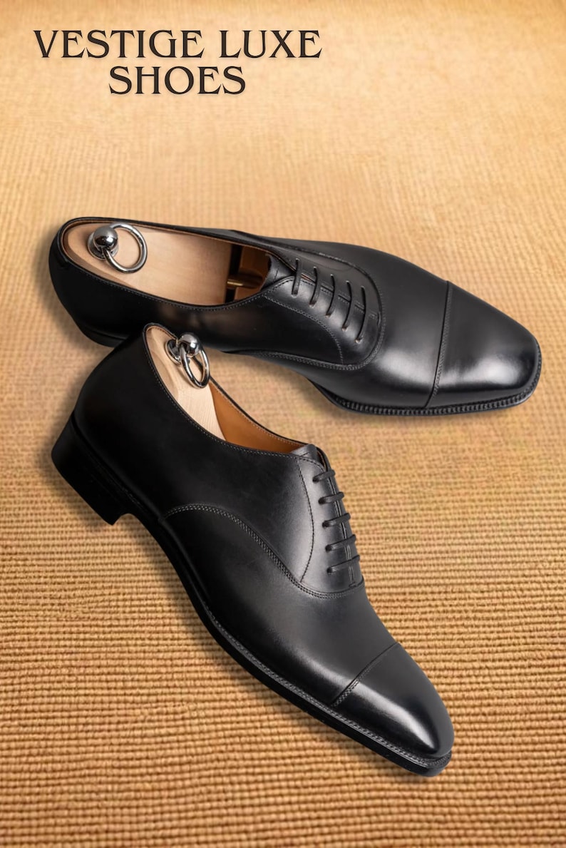 Puede incluir: Dos zapatos de vestir de cuero negro con hormas de madera. Los zapatos est&aacute;n atados con cordones y tienen un dise&ntilde;o de puntera. El texto "VESTIGE LUXE SHOES" est&aacute; en la parte superior de la imagen.