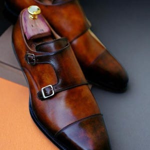 May include: A pair of brown leather monk strap shoes with a glossy finish. The shoes have a double buckle closure and are displayed with wooden shoe trees. The text "VESTIGE LUXE SHOES" is visible.