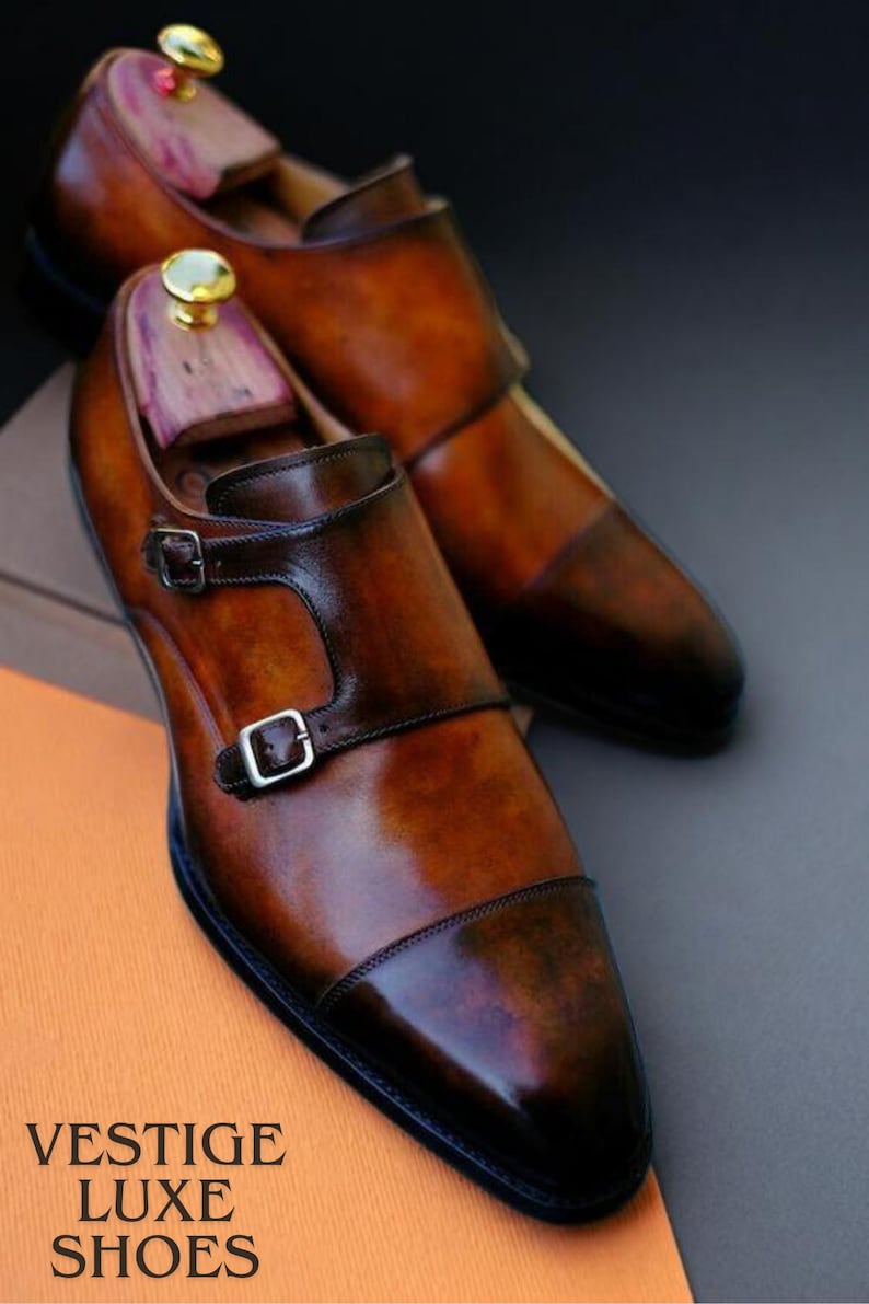 May include: A pair of brown leather monk strap shoes with a glossy finish. The shoes have a double buckle closure and are displayed with wooden shoe trees. The text "VESTIGE LUXE SHOES" is visible.