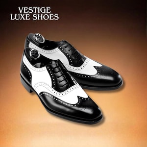 Può includere: Un paio di scarpe eleganti in pelle bicolore bianca e nera. Le scarpe presentano un classico design a coda di rondine con fori decorativi e chiusura con lacci. Il testo "VESTIGE LUXE SHOES" è in alto.