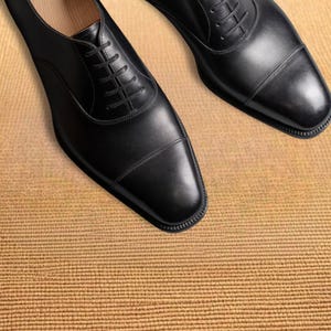 Puede incluir: Un par de zapatos de vestir de cuero negro con un dise&ntilde;o Oxford cl&aacute;sico. Los zapatos tienen cierre de cordones, punta redondeada y acabado pulido. Tambi&eacute;n se muestra el texto "VESTIGE LUXE SHOES".