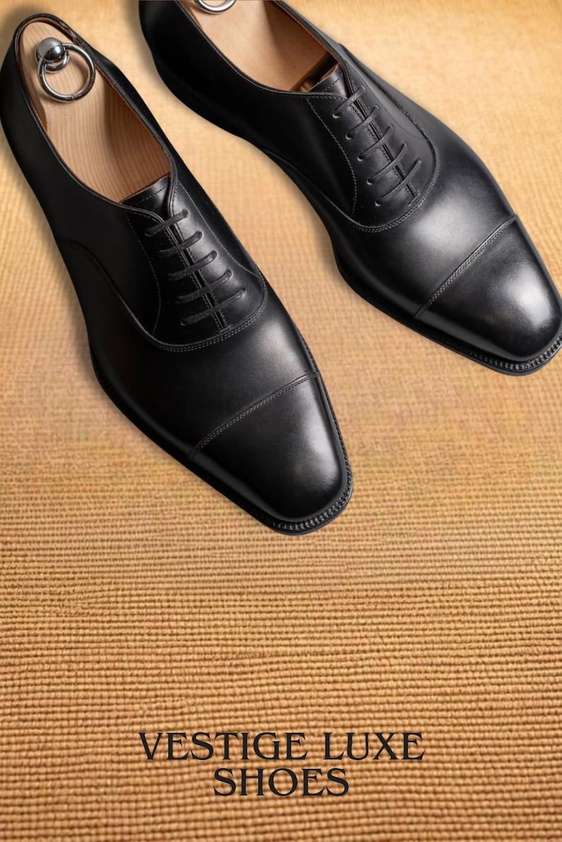 Puede incluir: Un par de zapatos de vestir de cuero negro con un dise&ntilde;o Oxford cl&aacute;sico. Los zapatos tienen cierre de cordones, punta redondeada y acabado pulido. Tambi&eacute;n se muestra el texto "VESTIGE LUXE SHOES".