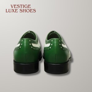May include: A pair of green and white wingtip shoes with black heels and soles. The text VESTIGE LUXE SHOES is printed in dark red in the upper left corner. The shoes feature decorative brogue perforations along the leather panels.