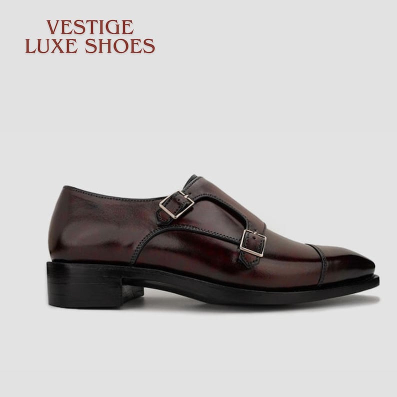 May include: Burgundy leather double monk strap shoe with silver buckles. The shoe has a black sole and heel. The text "VESTIGE LUXE SHOES" is at the top of the image.