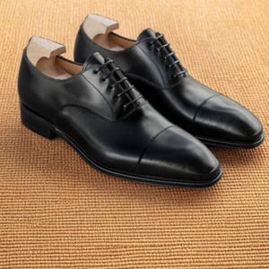 Puede incluir: Un par de zapatos de vestir de cuero negro con cordones, exhibidos sobre una superficie marr&oacute;n texturizada. Los zapatos tienen un dise&ntilde;o cl&aacute;sico con puntera y est&aacute;n etiquetados como "VESTIGE LUXE SHOES".