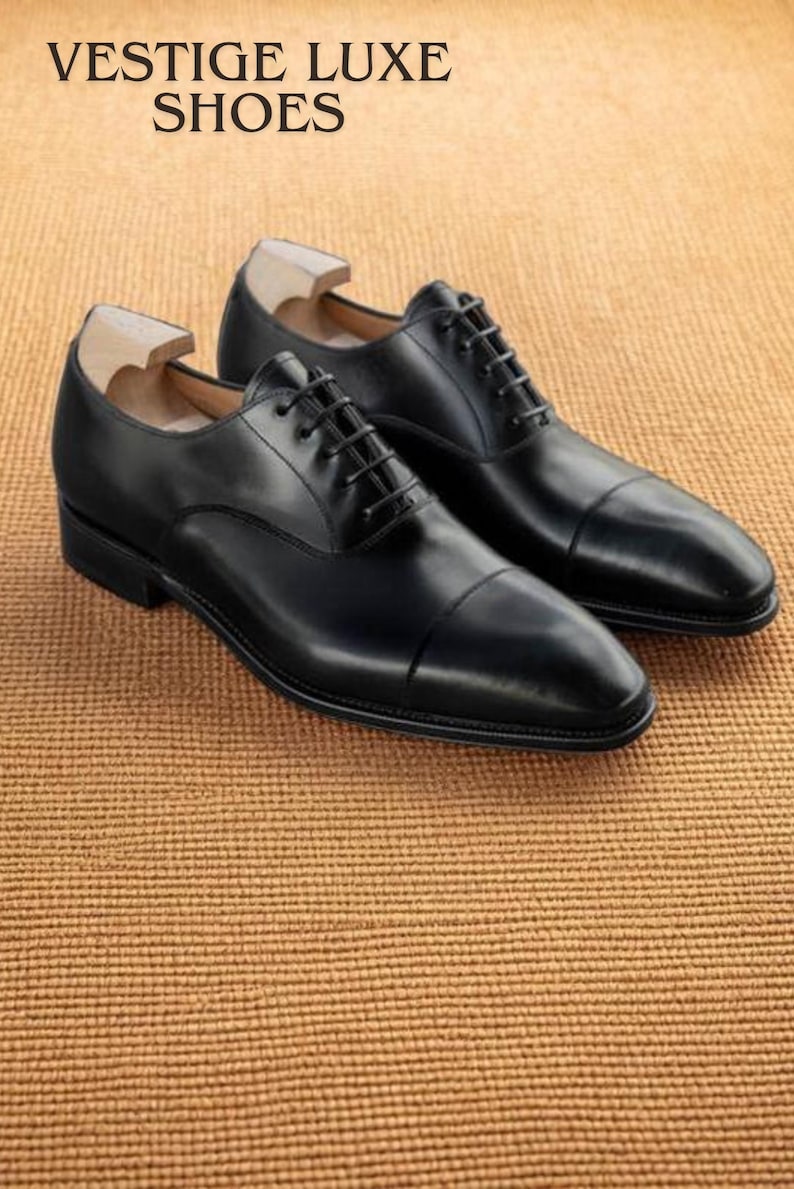 Puede incluir: Un par de zapatos de vestir de cuero negro con cordones, exhibidos sobre una superficie marr&oacute;n texturizada. Los zapatos tienen un dise&ntilde;o cl&aacute;sico con puntera y est&aacute;n etiquetados como "VESTIGE LUXE SHOES".