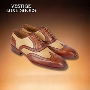 Könnte beinhalten: Zweifarbige Lederschuhe in Braun und Beige. Die Schuhe haben ein Flügelspitzendesign mit dekorativen Perforationen und braunen Schnürsenkeln. Der Text "VESTIGE LUXE SHOES" steht oben.