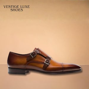 May include: Brown leather double-monk strap shoe with a pointed toe. The shoe features two silver buckles and a dark brown sole. The text "VESTIGE LUXE SHOES" is visible in the upper left corner.
