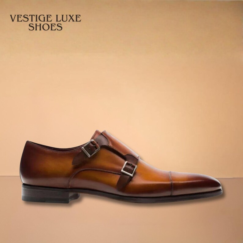 May include: Brown leather double-monk strap shoe with a pointed toe. The shoe features two silver buckles and a dark brown sole. The text "VESTIGE LUXE SHOES" is visible in the upper left corner.