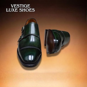 Pode incluir: Sapatos de couro verde com fivela dupla e fivelas prateadas. Os sapatos têm um acabamento brilhante e um design clássico. O texto "VESTIGE LUXE SHOES" está na parte superior da imagem.