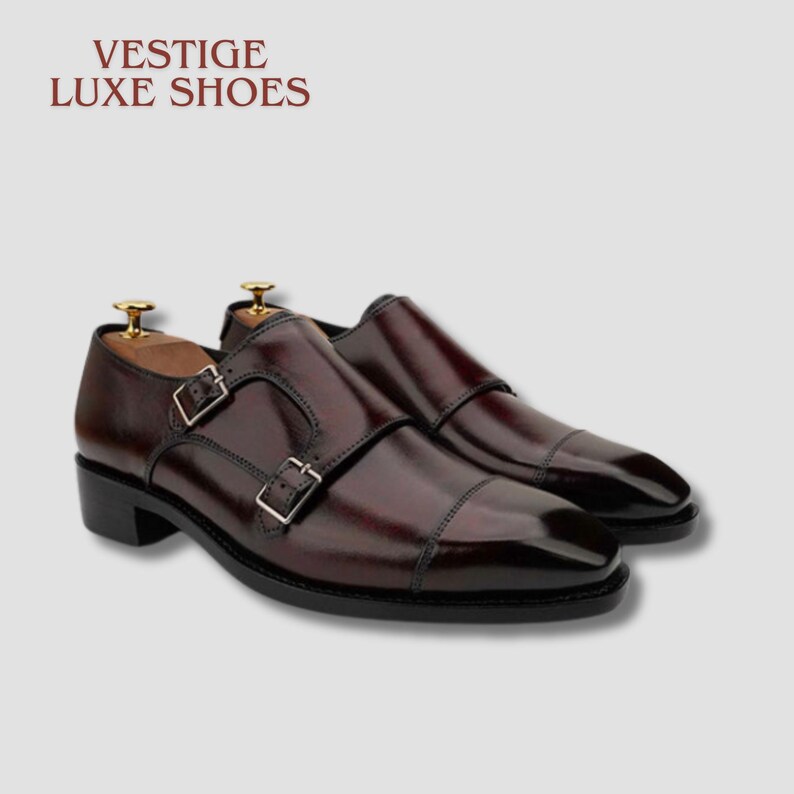 May include: A pair of burgundy leather double monk strap shoes with silver buckles. The shoes have a polished finish and a classic design. The text "VESTIGE LUXE SHOES" is at the top of the image.