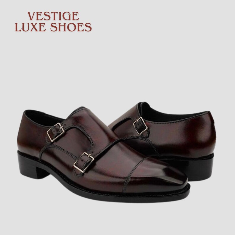 May include: A pair of burgundy leather double monk strap shoes with silver buckles. The shoes have a polished finish and a classic design. The text "VESTIGE LUXE SHOES" is at the top of the image.