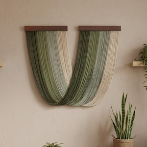 May include: A wall hanging with a wooden top and cascading strands in shades of green and beige. The strands are arranged in a curved, flowing design. The wall hanging is mounted on a neutral-colored wall, with plants in the foreground.