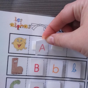 Puede incluir: Juego educativo de aprendizaje del alfabeto con tarjetas de letras. La imagen muestra una mano colocando la letra "A" en una tarjeta correspondiente. Se ven otras letras e ilustraciones, como "B", "C" y "D". El juego está diseñado para la educación infantil.
