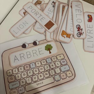 Peut inclure: Ensemble de cartes éducatives et un outil d'apprentissage en forme de clavier. Les cartes présentent des mots comme "LION" et "PORTE" avec des illustrations correspondantes. Le clavier affiche le mot "ARBRE" en haut, avec une disposition AZERTY complète.