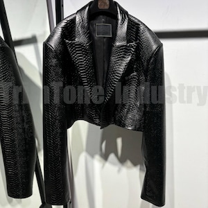 Może przedstawiać: Czarny, krótki żakiet z teksturowanym wzorem przypominającym skórę węża. Kurtka ma klapy z wycięciami, długie rękawy i dopasowany krój. Tekst "Tri Tone Industry" jest widoczny w tle.