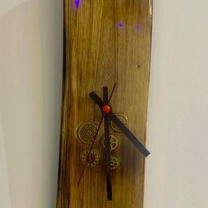 May include: Wooden wall clock with a natural wood grain finish. The clock features gold numerals at the 12 and 6 positions, with decorative gears and black clock hands. The clock is rectangular and mounted on a white wall.