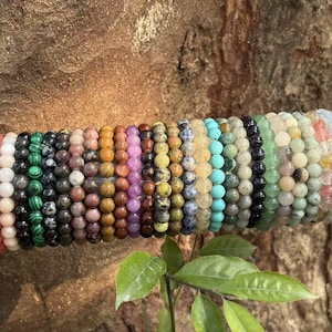 May include: A collection of beaded bracelets in various colors, including pink, white, green, brown, orange, and blue. The bracelets are made of small, round beads and are displayed on a gray cylindrical support.