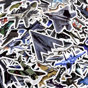 Puede incluir: Una colección de pegatinas de aviones con varios diseños de aeronaves. Las pegatinas muestran una variedad de aviones militares y civiles en diferentes colores y estilos, incluyendo aviones de combate y aviones de hélice.