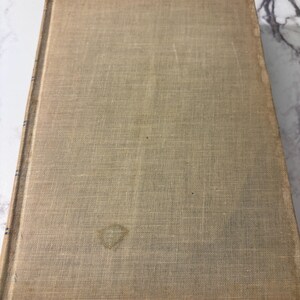 May include: A vintage book with a beige linen cover. The cover shows signs of wear, including a water stain and discoloration along the edges. The book is closed and resting on a marble surface.