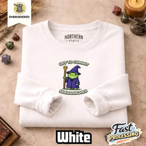 May include: White long-sleeved sweatshirt featuring a cartoon frog wizard. The frog is dressed in a purple robe and hat, holding a staff, with the words "OFF TO COMMIT SHENANIGANS". The sweatshirt has the word "WHITE" at the bottom.