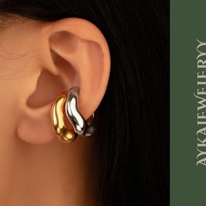 Puede incluir: Primer plano de una oreja adornada con dos pendientes curvos apilados. El pendiente inferior es dorado, mientras que el superior es plateado. Los pendientes son lisos y brillantes. La imagen incluye el texto "AYKAJEWELERYY" en una fuente estilizada.