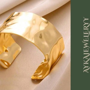 Könnte beinhalten: Ein goldfarbenes Armreif-Armband mit strukturierter, gehammerter Oberfläche. Das breite, offene Design hat eine geschwungene Form und wird auf einem neutralen Stoff präsentiert. Der Text "AYKAJEWELRY" ist auf grünem Hintergrund zu sehen.