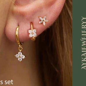 Könnte beinhalten: Ein Set aus drei goldfarbenen Ohrringen, darunter ein Paar Creolen mit einem hängenden Blumenanhänger, ein Paar kleine Creolen mit einem Blumenanhänger und ein Blumenstecker. Der Text "3pcs set" ist sichtbar.