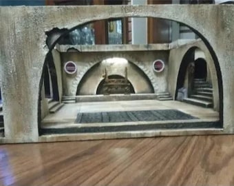 May include: A detailed diorama of a building interior with arched doorways and a central courtyard. The structure is painted in shades of beige and brown, with a dark wooden door on the right. The interior features stairs and a decorative element.