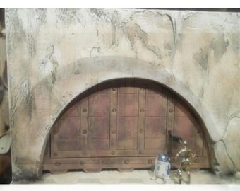 May include: A detailed diorama featuring a weathered, arched doorway with a wooden door. The scene includes R2-D2 and C-3PO figurines, set against a textured, tan-colored wall, evoking a desert environment.