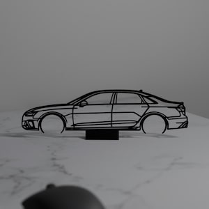 May include: A black line art car silhouette on a small black stand. The car is a sedan style with visible details like the wheels and windows. The image is set on a white surface with a computer monitor and a coffee mug in the background.
