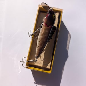 May include: A vintage fishing lure in a yellow box. The lure is brown with red accents and silver hooks. The lure has a propeller and is designed to mimic a small fish. The box is rectangular and has a yellow exterior.