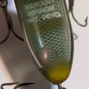 Könnte beinhalten: Ein Vintage-Angelköder mit einem transparenten, grünen und gelben Körper. Der Köder hat einen silbernen Haken und der Text "MERCURY MINNOW PAT. PEND." ist sichtbar. Der Köder hat ein silbernes Gewicht im Inneren und ein Netzmuster.
