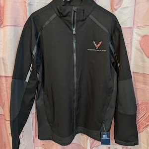 Puede incluir: Chaqueta negra con cremallera con la palabra "CORVETTE" y un logotipo rojo y plateado. La chaqueta tiene un cuello alto, detalles en gris y una cremallera completa. La chaqueta está colgada en una percha de madera.