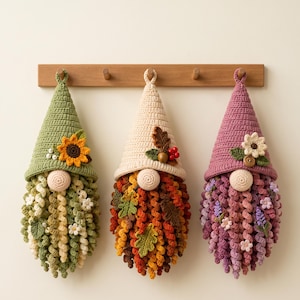 May include: Three hanging crochet gnomes with pointed hats. The left gnome has a green hat with a sunflower, the center has a cream hat with an acorn and leaves, and the right has a mauve hat with a flower. Each has a curly beard and nose.