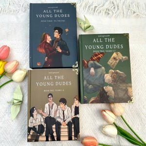 May include: Three hardcover books titled All The Young Dudes, featuring illustrated covers. Book one is brown, book two is green, and book three is dark blue. Each cover displays character artwork, gold corner accents, and printed text.