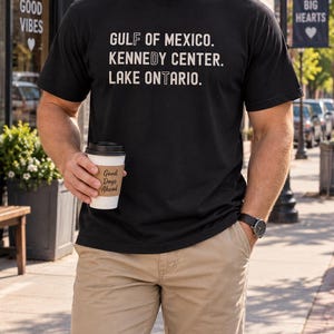 Könnte beinhalten: Ein schwarzes kurzärmeliges T-Shirt mit dem weißen Text GULF OF MEXICO. KENNEDY CENTER. LAKE ONTARIO. Das Shirt wird mit khakifarbenen Shorts und einer Armbanduhr kombiniert, während ein weißer Kaffeebecher mit der Aufschrift Good Days Ahead gehalten wird.