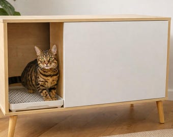 May include: A contemporary cat litter box cabinet in light wood with a white sliding door. A brown tabby cat is inside the open section, which has a litter tray. The cabinet has angled wooden legs and is on a wooden floor.