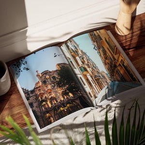 May include: Open magazine featuring architectural photography of buildings with ornate facades and balconies. The pages are spread on a wooden surface, with a gray ceramic cup and a decorative wooden object nearby. Green plants are in the foreground.