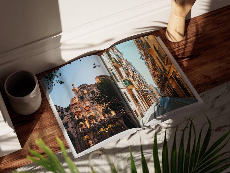 May include: Open magazine featuring architectural photography of buildings with ornate facades and balconies. The pages are spread on a wooden surface, with a gray ceramic cup and a decorative wooden object nearby. Green plants are in the foreground.