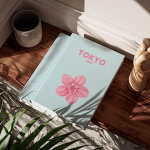 May include: A light blue journal with the word "TOKYO" and the year "2025" in pink. A pink cherry blossom is on the cover. The journal is on a wooden surface, next to a small, gray ceramic pot and a wooden hand sculpture.