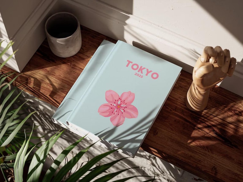 May include: A light blue journal with the word "TOKYO" and the year "2025" in pink. A pink cherry blossom is on the cover. The journal is on a wooden surface, next to a small, gray ceramic pot and a wooden hand sculpture.