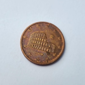 Peut inclure: Une pièce de cinq centimes d'euro italienne de couleur cuivre représentant le Colisée en relief. La pièce affiche l'année 2002, les lettres IR et un cercle de douze étoiles. La surface présente des signes d'oxydation et d'usure.
