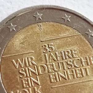 May include: A two-tone silver and gold commemorative 2-euro coin featuring the text 35 JAHRE WIR SIND EIN VOLK JAHRE EINHEIT, the letter D, and the year 2025. The coin has a metallic border with stars and a central gold-colored circular design.