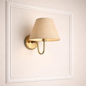 May include: A wall-mounted sconce with a beige fabric shade and a brass-colored metal base. The sconce is mounted on a white wall within a white framed border. The lamp's design is simple and elegant, suitable for various interior styles.